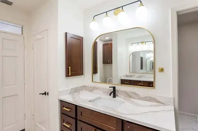 Bathroom vanity with a dark wood cabinet, white quartz top, arched brass mirror and a three-globe light